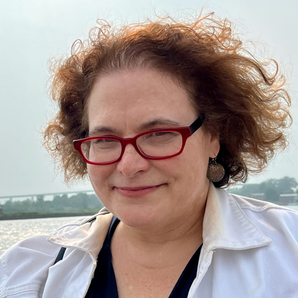 Headshot of a woman with dark curly hair and glasses. There is a skyline and lake in the background.